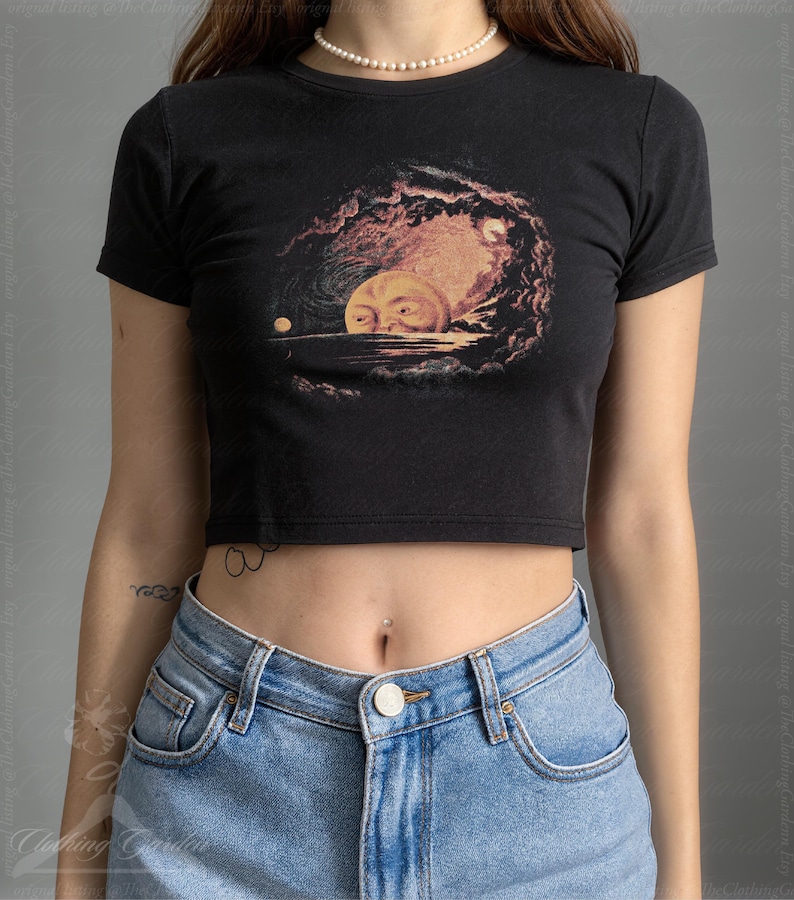 May include: Black crop top featuring a celestial design with a large, smiling moon and a smaller planet. The graphic is in shades of orange, pink, and yellow against a dark background. The top has short sleeves and a crew neck.
