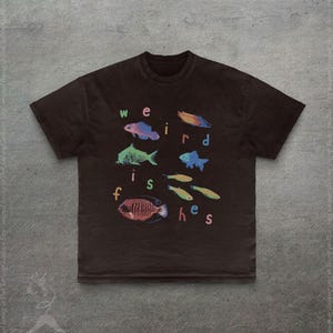 Puede incluir: Una camiseta marr&oacute;n oscuro con la frase "weird fishes" en letras coloridas. La camiseta muestra varias ilustraciones de peces coloridos, incluyendo peces azules, verdes y rojos. El dise&ntilde;o est&aacute; centrado en la parte delantera de la camiseta.