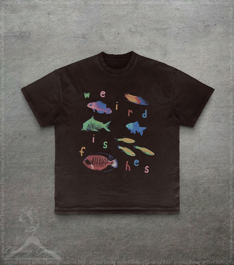 Puede incluir: Una camiseta marr&oacute;n oscuro con la frase "weird fishes" en letras coloridas. La camiseta muestra varias ilustraciones de peces coloridos, incluyendo peces azules, verdes y rojos. El dise&ntilde;o est&aacute; centrado en la parte delantera de la camiseta.