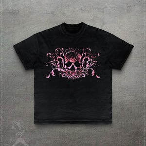 May include: Black t-shirt featuring a pink skull design with floral accents. The skull graphic is centered on the front of the shirt. The shirt has short sleeves and a crew neck.