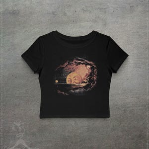 May include: Black crop top featuring a surreal illustration of a moon with a face, set against a dark, cloudy background. The artwork includes a small, round celestial body and a body of water. The shirt is made of a soft material.