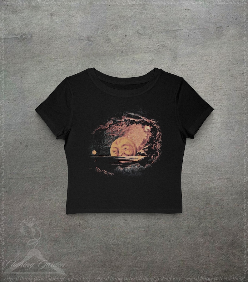 May include: Black crop top featuring a surreal illustration of a moon with a face, set against a dark, cloudy background. The artwork includes a small, round celestial body and a body of water. The shirt is made of a soft material.
