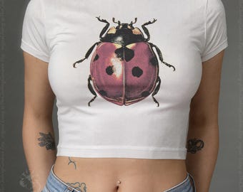 May include: A white short-sleeved crop top featuring a large, detailed graphic print of a ladybug on the center chest. The ladybug has a pinkish-red shell with distinct black spots and a black head and thorax. The shirt has a crew neckline.
