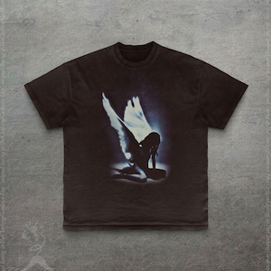 Pode incluir: Camiseta marrom escuro com um gr&aacute;fico desgastado de um anjo ajoelhado com grandes asas brancas e um halo azul. A imagem est&aacute; centrada na camiseta. A camiseta tem mangas curtas e gola redonda.