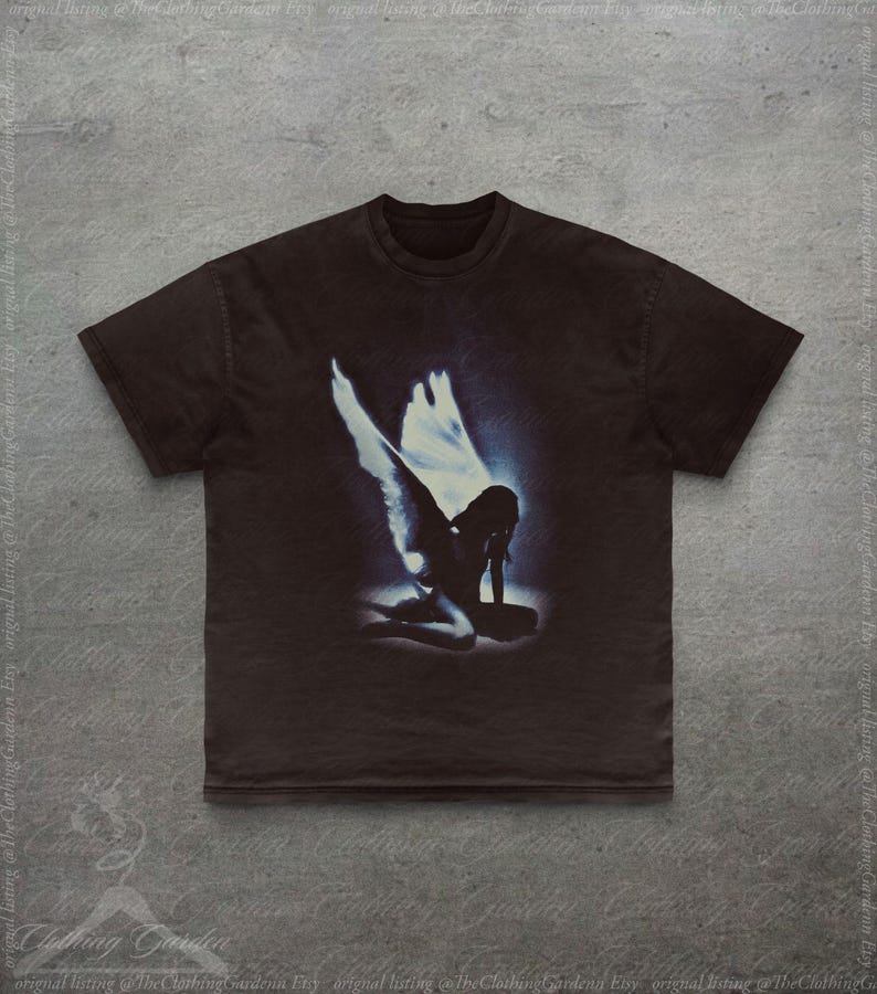 Pode incluir: Camiseta marrom escuro com um gr&aacute;fico desgastado de um anjo ajoelhado com grandes asas brancas e um halo azul. A imagem est&aacute; centrada na camiseta. A camiseta tem mangas curtas e gola redonda.