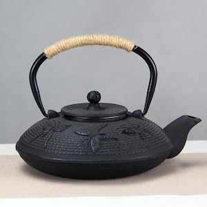 Puede incluir: Tetera de hierro fundido negro con diseño de libélula. La tetera tiene un asa negra curvada envuelta con cuerda de color natural. La tapa tiene un pequeño pomo. El pico está en el lado derecho.