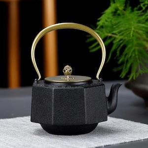 May include: Black cast iron teapot with a gold-coloured handle and lid. The teapot has an octagonal body and a curved spout. The lid is adorned with a decorative knob. The teapot is sitting on a grey surface.