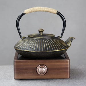 May include: A black and gold cast iron teapot with a rope-wrapped handle and a matching lid. The teapot sits on a wooden base with a copper-colored knob, ideal for brewing tea. The teapot has a traditional design.