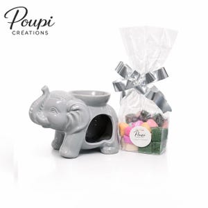 Peut inclure: Un brûleur d'huile en forme d'éléphant gris avec un bol sur le dessus et une grande ouverture latérale. Un sac transparent de fondants parfumés colorés est noué avec un ruban argenté. La marque "Poupi Créations" est visible.