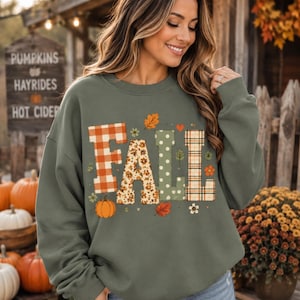 May include: An olive green crewneck sweatshirt featuring the word FALL printed in large letters. Each letter has a unique pattern: orange plaid, floral print, green polka dots, and tan plaid. Small autumn-themed icons like leaves and pumpkins surround the text.