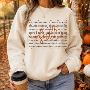 May include: A cream-colored crewneck sweatshirt featuring a list of autumn-themed words printed in black, including BONFTRES, FLANNELS, APPLE PICKING, and more, with the word fall in large orange script and small orange hearts between the words.