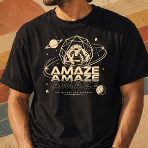 May include: A black short-sleeve t-shirt featuring a cream-colored graphic of a geometric sphere containing a crab-like creature, surrounded by planets and stars. The text reads AMAZE AMAZE AMAZE and BEYOND ORDINARY in a stylized font.
