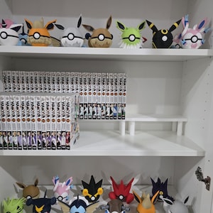 Może przedstawiać: Biała szafa z kilkoma półkami prezentuje kolekcję pluszowych postaci Pokémon i tomów mangi. Pluszaki, w tym ewolucje Eevee, są ułożone na górnej i dolnej półce. Seria mangi "Haikyu!!" jest widoczna.