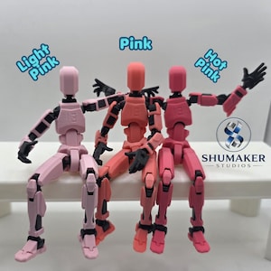 Może przedstawiać: Trzy przegubowe figurki akcji z czarnymi stawami, oznaczone jako Light Pink, Pink i Hot Pink. Figurki są w pozycji siedzącej. Logo Shumaker Studios jest widoczne po prawej stronie, wraz z tekstem Light Pink, Pink i Hot Pink.