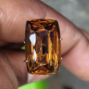 Può includere: Un anello con una grande gemma color ambra a taglio rettangolare. La gemma è tenuta in posizione da due griffe d'argento. L'anello è tenuto in mano su uno sfondo sfocato.