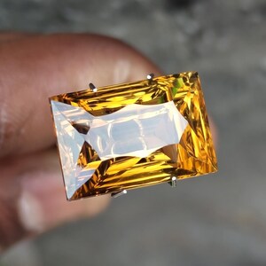 Puede incluir: Una gema grande, rectangular y facetada con un vibrante tono amarillo-naranja. La gema se sujeta entre dos puntas plateadas, mostrando su claridad y brillo. El corte de la piedra crea múltiples superficies reflectantes, realzando su brillo.