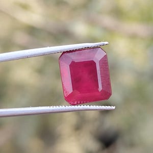 Könnte beinhalten: Ein leuchtender, rechteckig geschliffener Rubin, der von einer Pinzette gehalten wird. Der Edelstein hat eine tiefe, satte rote Farbe mit polierter Oberfläche. Der Hintergrund ist unscharf, wodurch die Klarheit und Brillanz des Edelsteins hervorgehoben wird.
