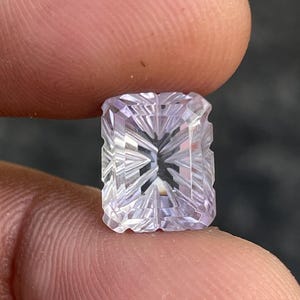 May include: A rectangular, light lavender gemstone featuring a complex, multi-faceted cut with sharp, geometric lines radiating from the center. The stone displays a translucent quality with a high-gloss finish, reflecting light across its surface.