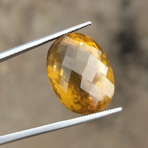 May include: A faceted, oval-shaped gemstone held by a pair of tweezers. The gem is a warm, golden-yellow color, with multiple facets that catch the light. The tweezers are silver-colored and have serrated edges.