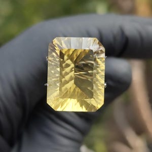 May include: A large, rectangular-cut, yellow gemstone. The gem is held against a blurred background, showcasing its facets and internal patterns. The stone's color ranges from pale to a rich, golden yellow.