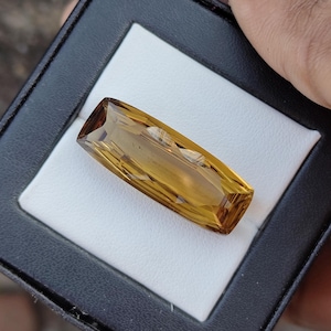 May include: A rectangular, golden-yellow gemstone with a faceted cut, displayed in a black case with a white interior. The gemstone has a warm, amber hue and is held by a hand.