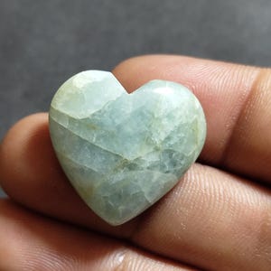 May include: A heart-shaped polished gemstone with a pale mint green and translucent appearance. The stone features visible internal fractures and natural veining throughout its surface, creating a smooth, glossy texture with subtle color variations.