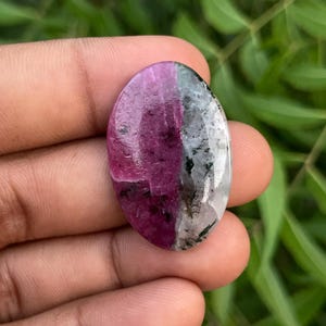 Puede incluir: Una piedra preciosa en cabujón de forma ovalada con un patrón de color dividido. Una mitad es de color rosa magenta intenso con inclusiones oscuras, mientras que la otra mitad es de color blanco translúcido con motas minerales negras.