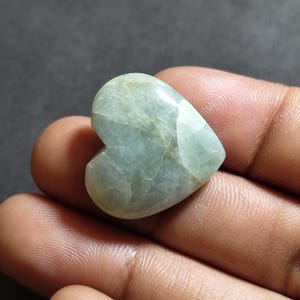 May include: A polished heart-shaped stone with a smooth, glossy surface. The stone features a pale, translucent mint green color with subtle internal veining and cloudy white inclusions throughout its structure, creating a natural marbled appearance.