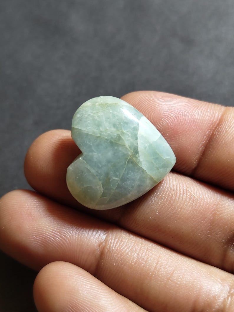 May include: A polished heart-shaped stone with a smooth, glossy surface. The stone features a pale, translucent mint green color with subtle internal veining and cloudy white inclusions throughout its structure, creating a natural marbled appearance.