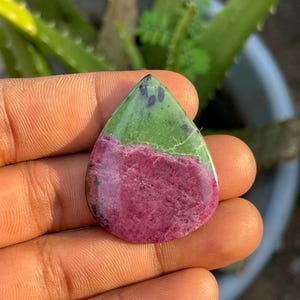 Puede incluir: Una piedra preciosa de rubí zoisita pulida en forma de lágrima, con una clara división de color entre una sección inferior de color rosa magenta vibrante y una sección superior de color verde lima, con pequeñas inclusiones oscuras.