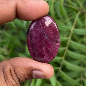 Puede incluir: Una piedra preciosa pulida de forma ovalada con una paleta de colores magenta intenso y rojo oscuro. La piedra presenta una superficie lisa y reflectante con texturas cristalinas internas visibles e inclusiones naturales en todo su cuerpo sólido.