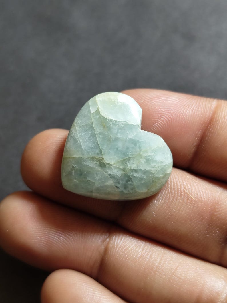 May include: A heart-shaped polished gemstone with a pale mint green and white marbled appearance. The stone features a smooth, glossy surface with visible internal fractures and natural veining patterns throughout its translucent structure.