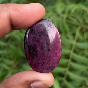 Può includere: Una pietra preziosa lucida di forma ovale con una profonda tonalità magenta e viola, caratterizzata da inclusioni scure e irregolari su tutta la superficie liscia e riflettente. La pietra presenta una texture naturale screziata con varie sfumature.