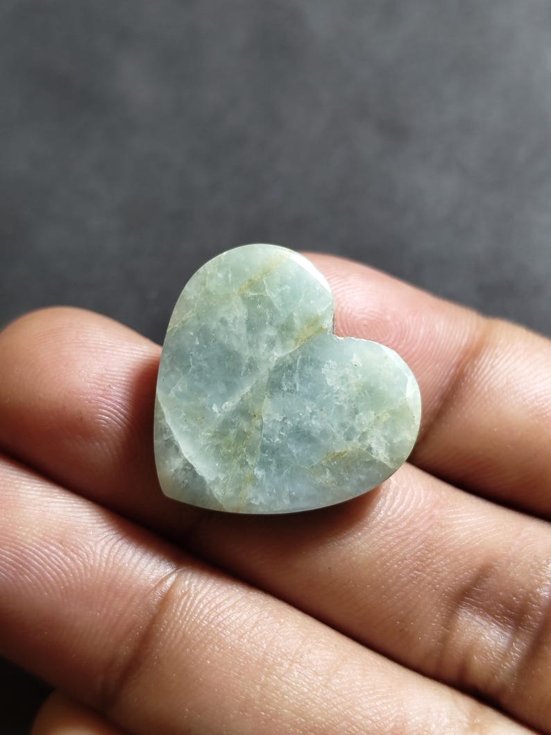 May include: A heart-shaped polished stone with a pale mint green color and translucent quality. The surface features visible internal fractures and subtle golden-brown veining throughout the smooth, rounded edges of the gemstone.