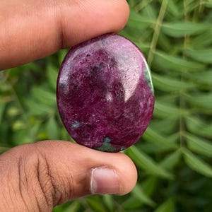 Puede incluir: Una piedra preciosa pulida de forma ovalada que presenta una superficie de color magenta intenso y rojo oscuro con inclusiones visibles de depósitos minerales de color verde oscuro, mostrando una textura suave y reflectante con un patrón natural.