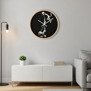 May include: Round wall clock with a black face and a light wood frame. The clock features a black and white image of a person holding a puppet. The clock hands are white.