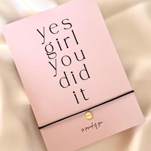 Könnte beinhalten: Eine rosa Karte mit den Worten "yes girl you did it" in schwarzer Schrift. Ein schwarzes Kordelarmband mit einem kleinen goldenen Anhänger ist an der Karte befestigt. Der Text "So proud of you" steht unten.
