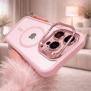May include: A pink phone case with a clear back and a metallic ring holder. The case has a built-in magnetic circle with an Apple logo. The ring holder is attached to the camera area and has the word "SUPPORT" engraved on it.