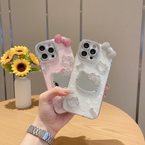 May include: Two decorative phone cases, one pink and one white, with Hello Kitty designs. Each case has a bow and is embellished with rhinestones. The cases are held in front of a vase of sunflowers.