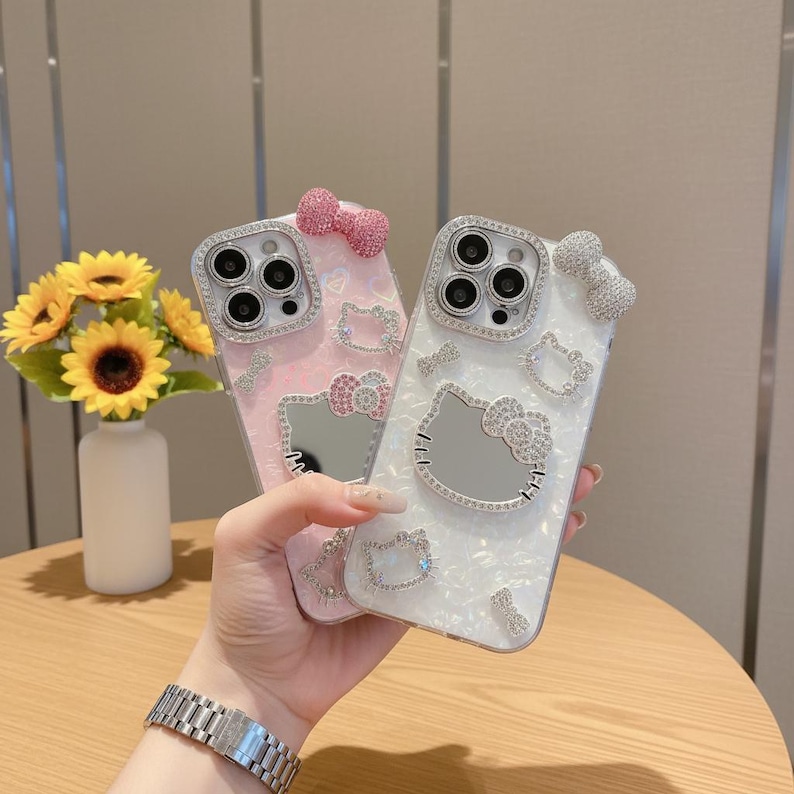 May include: Two decorative phone cases, one pink and one white, with Hello Kitty designs. Each case has a bow and is embellished with rhinestones. The cases are held in front of a vase of sunflowers.