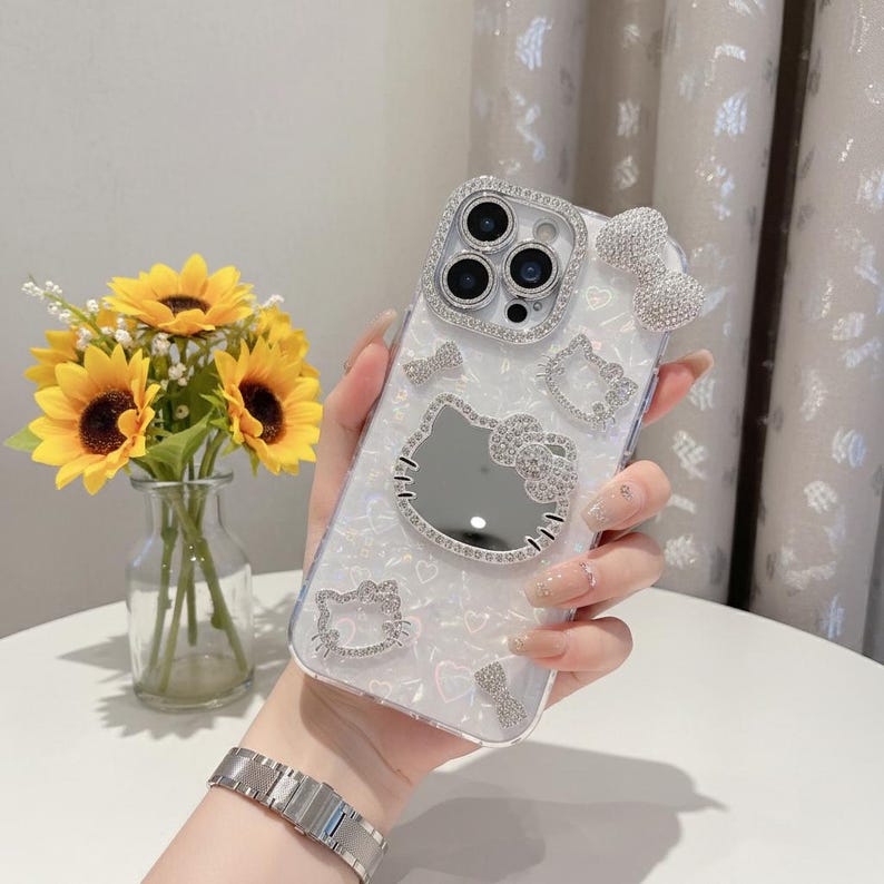 May include: A clear phone case with a pearlescent finish, decorated with sparkling rhinestones. The case features a mirrored Hello Kitty design, bow accents, and a rhinestone-encrusted camera frame. A bouquet of sunflowers is in the background.