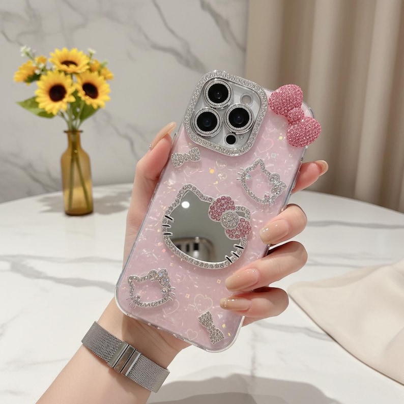 May include: Pink phone case with a Hello Kitty design. The case has a mirror, a pink bow, and rhinestone accents. The camera lens is surrounded by a silver rhinestone border. The case is held in a hand.