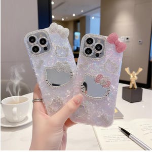 May include: Two iridescent phone cases with Hello Kitty designs. Each case features a sparkling bow, a mirrored Hello Kitty face, and rhinestone accents. The cases are designed to fit a smartphone and offer a stylish, protective accessory.