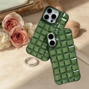May include: Two green phone cases with a woven pattern are displayed. A silver ring sits in front of the cases. The cases have camera cutouts and are on a light-colored surface. Pink and white roses are in the background.