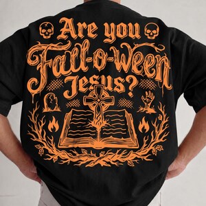 May include: A black t-shirt featuring an orange graphic on the back. The text reads "Are you Fall-o-ween Jesus?" above an illustration of an open book, a cross, a tombstone, a hand reaching from the ground, flames, and two skulls.