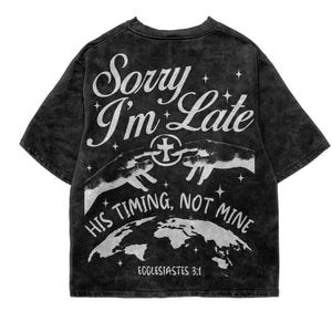 May include: Black acid wash t-shirt with white graphic text and illustrations. The text reads "Sorry I'm Late" above "His Timing, Not Mine" and "Ecclesiastes 3:1". The design includes hands, a cross, and a world map.