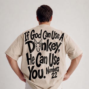 Puede incluir: Una camiseta beige de manga corta con texto negro en la espalda que dice: If God Can Use A Donkey, He Can Use You. Numbers 22. El diseño incluye un pequeño gráfico negro de la cabeza de un burro situado entre las palabras God y Donkey.