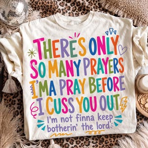 Puede incluir: Una camiseta de color crema con texto multicolor que dice: There's only so many prayers I'ma pray before I cuss you out. I'm not finna keep botherin' the lord. El diseño incluye pequeños elementos gráficos como un corazón, una estrella y un rayo.