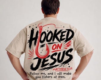 Peut inclure: T-shirt beige avec l'inscription "HOOKED ON JESUS" en noir et rouge, avec un motif de crochet de pêche rouge. Le texte "MATTHIEU 4:19" et un verset biblique sont également imprimés. C'est un vêtement décontracté.