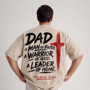 Peut inclure: Un t-shirt beige à manches courtes avec un texte noir style coup de pinceau indiquant DAD A MAN OF FAITH. A WARRIOR OF GOD. A LEADER OF HOME. et Proverbs 20:7 en rouge. Une grande croix rouge est placée à droite du texte au dos.